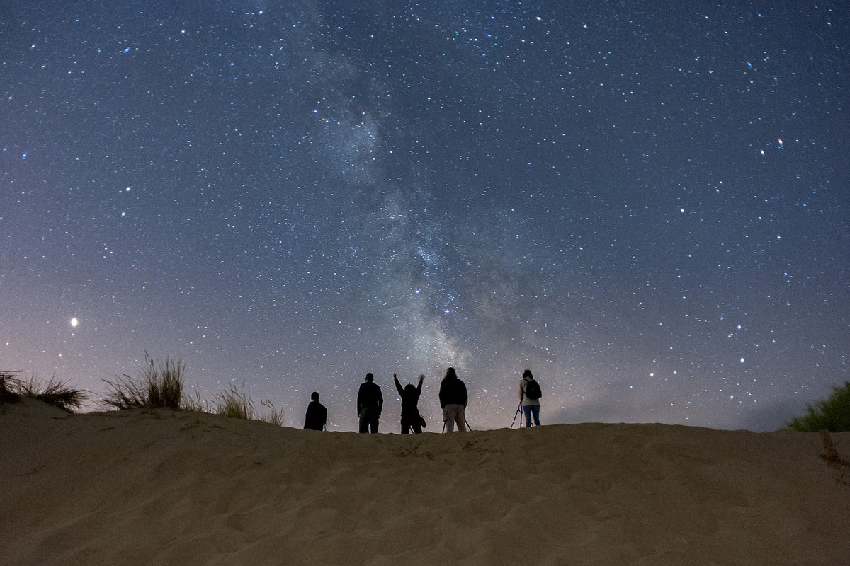 A group of five people standing on the beach looking up at a starry night sky and the milky way.